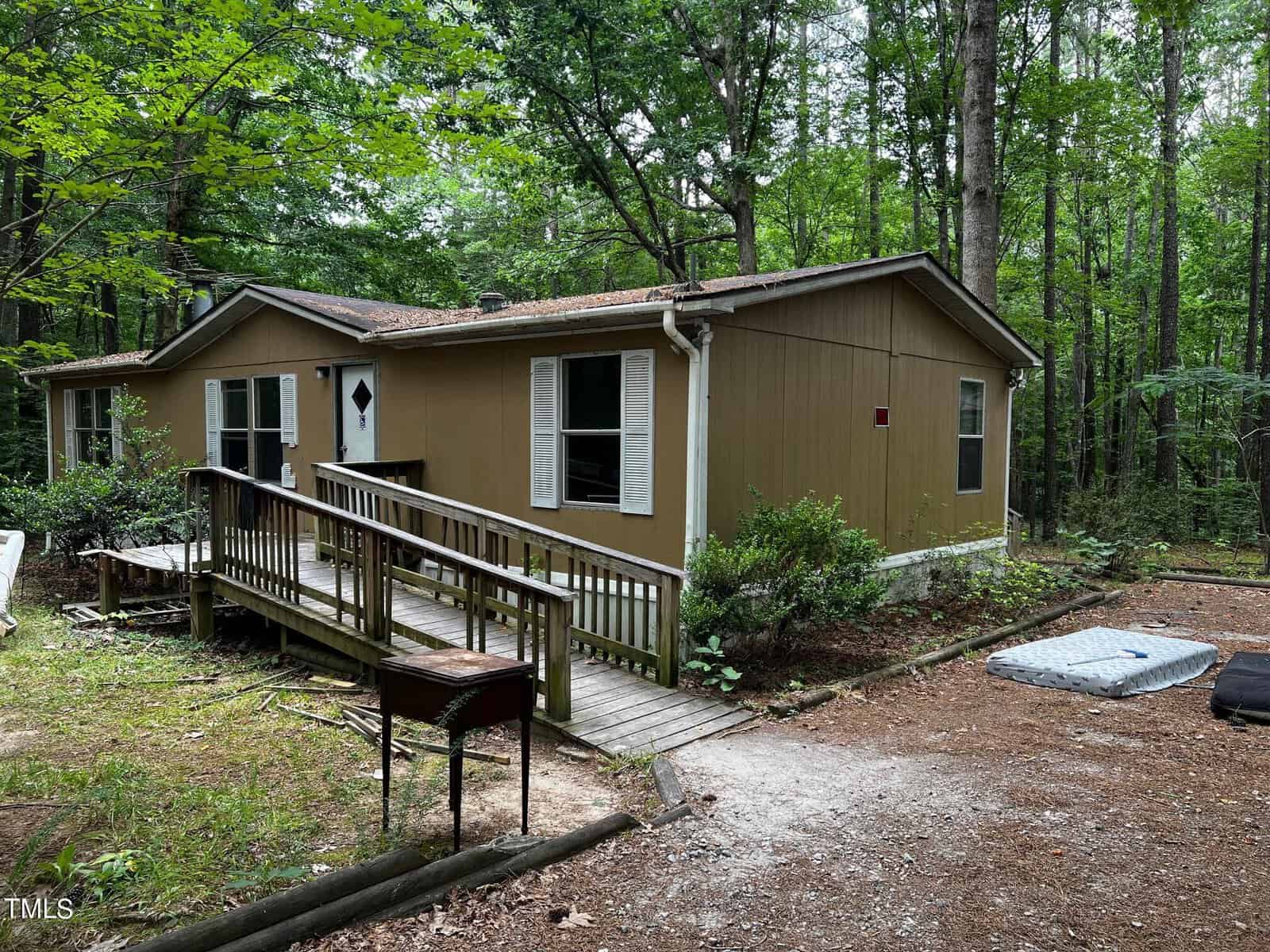 A small, brown, single-story house on 615 Shortleaf Court with white trim nestled in a wooded area of Wake Forest. The front entrance features a ramp leading up to a white door. Nearby, a black grill stands ready near the ramp amidst trees and scattered outdoor items.