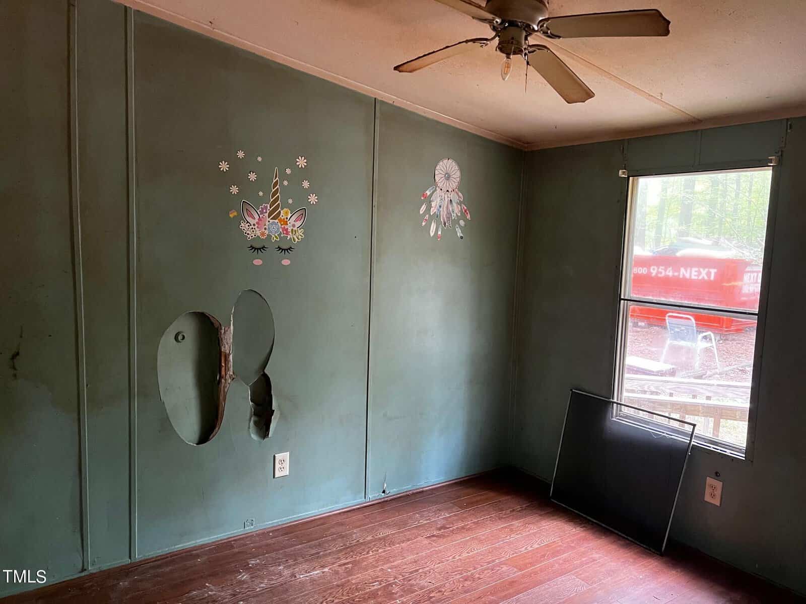 A small room with dark green walls, featuring wall art of butterflies and flowers. There's a hole in the wall, a ceiling fan, a window with a missing screen, and a hardwood floor. Outside, a red vehicle trailer is visible.