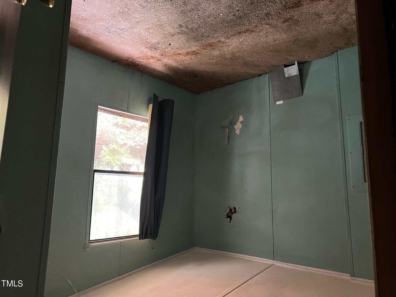 A small, empty room with pale green walls and a damaged ceiling. There's a window with a dark curtain on the left, some peeling paint, and a missing outlet cover. The room appears to be in need of renovation.