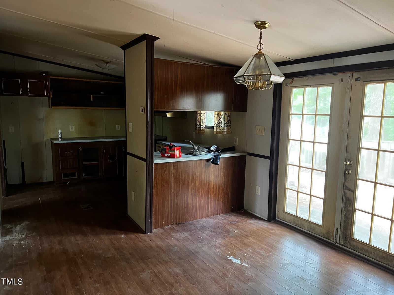 An empty room with a wooden floor, brown wood-paneled walls, and a kitchen counter. A chandelier hangs from the ceiling. French doors lead outside, and there's some clutter on the counter.