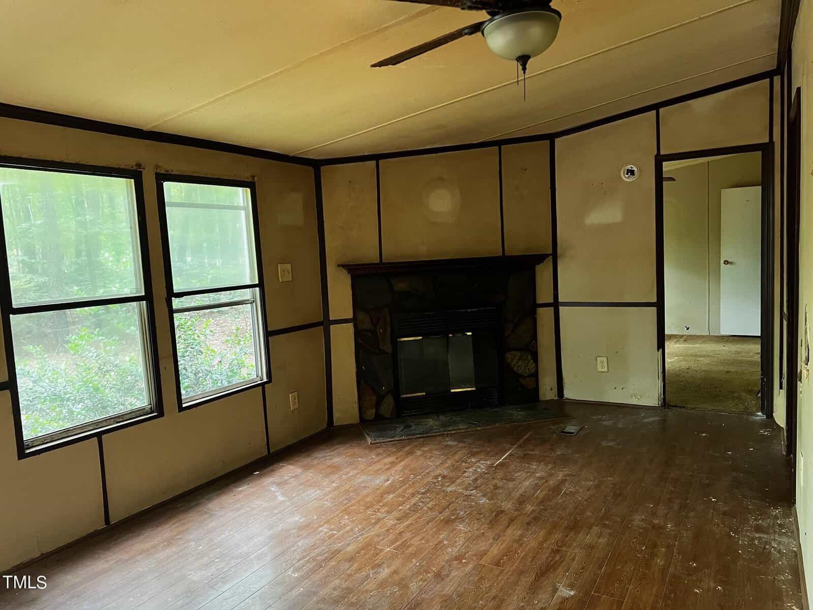 An empty room with wooden floors and a window. A fireplace is set into the wall, and a ceiling fan is above. The walls have wooden trim, and a doorway leads to another room with a closed door.