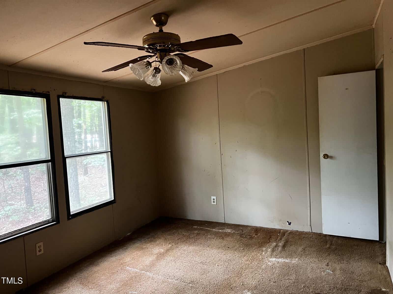 An empty room with beige walls, a ceiling fan with lights, and two rectangular windows. The floor is carpeted in brown, and a closed white door is visible on the right. Natural light enters through the windows, revealing a wooded area outside.