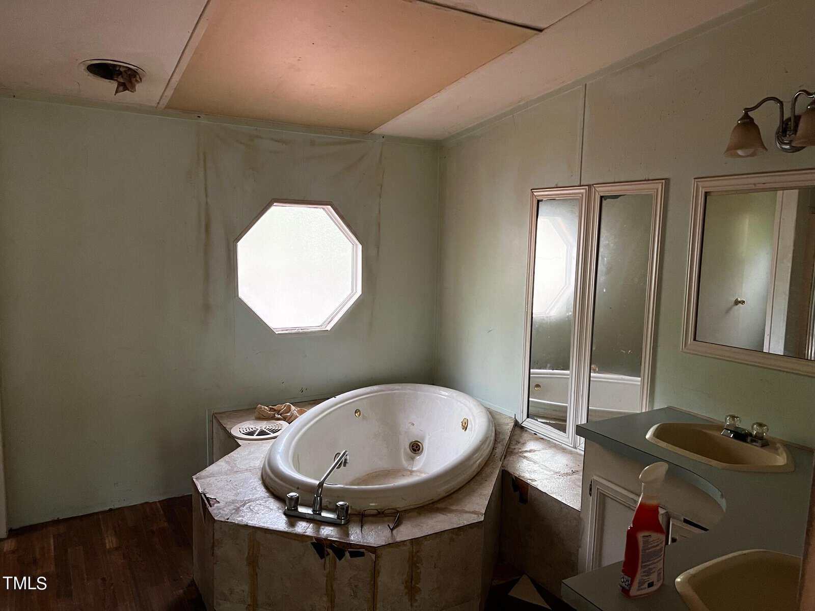 A bathroom with an octagonal window above a corner bathtub. The room has light green walls, a double sink with a large mirror, and a bottle of cleaner on the counter. The floor appears to be wood. The ceiling shows signs of wear.