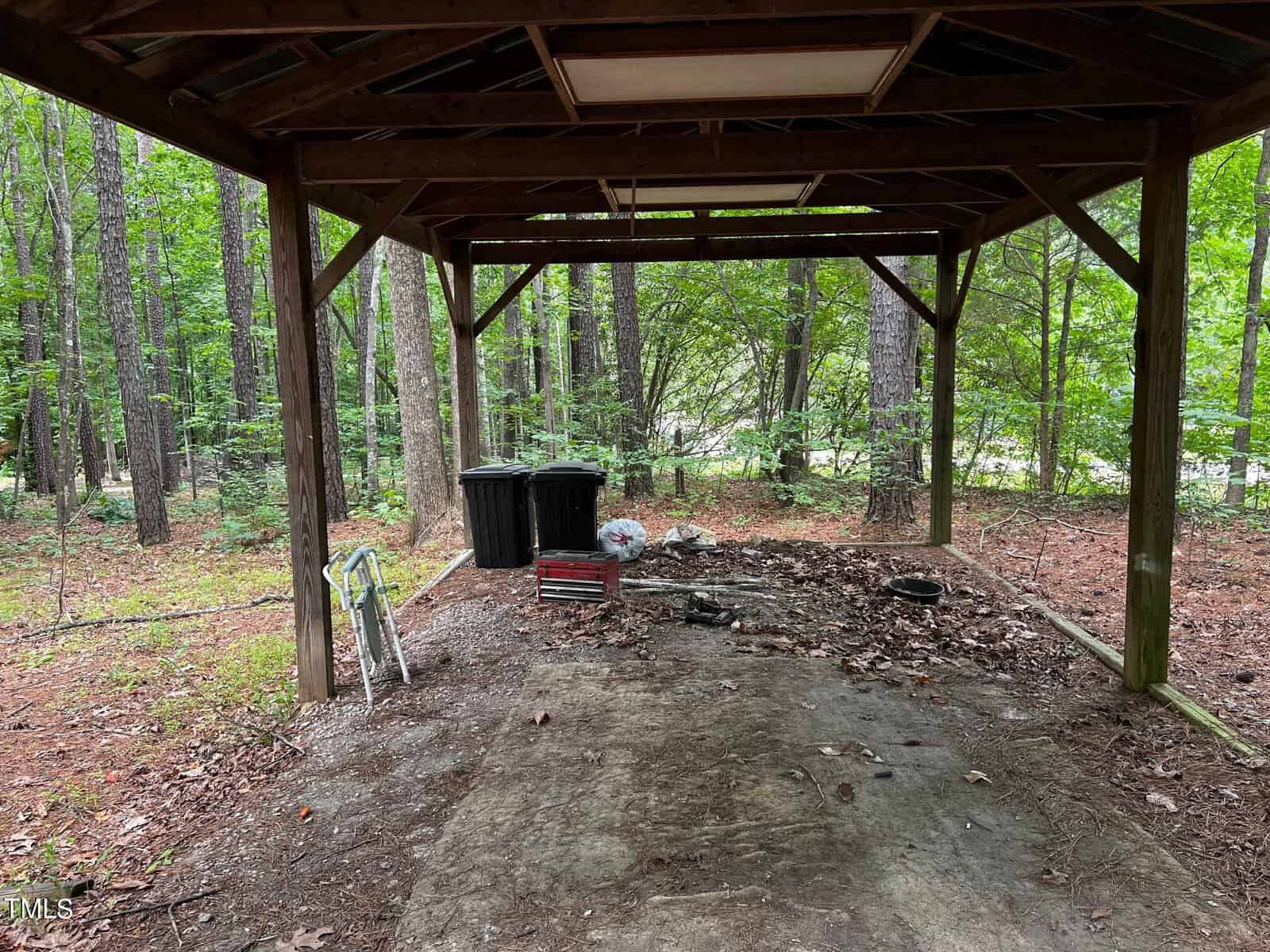A wooden canopy in a forest setting shelters various items, including three trash bins, a folding chair, a red toolbox, and other tools on a concrete slab. Tall trees and leafy undergrowth surround the structure.
