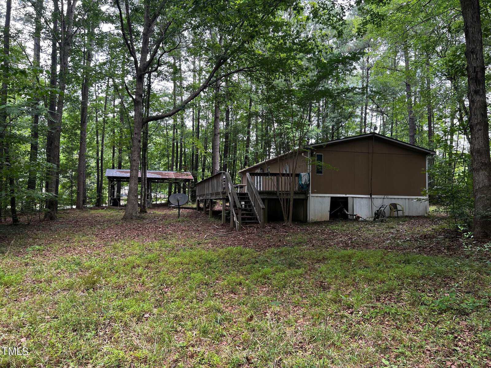 A brown mobile home is surrounded by tall trees, with a wooden deck extending from the front. A small dirt path leads up to the home. In the background, there is a metal-roofed structure nestled among the greenery.