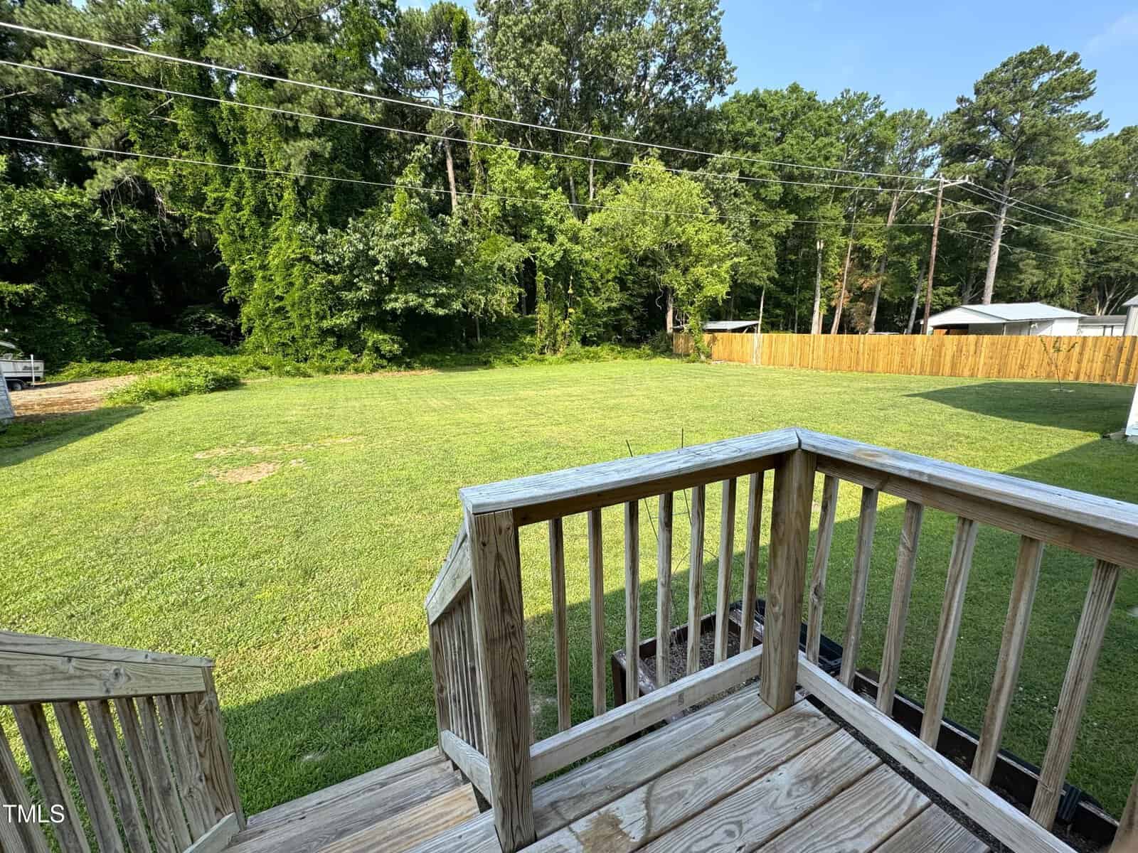 Wooden stairs lead down to a large, grassy backyard bordered by a wooden fence. The area is surrounded by lush trees under a clear blue sky.