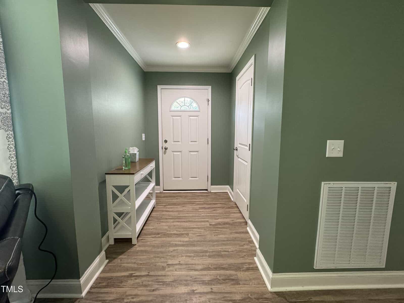 A hallway with green walls and wood flooring. It features a white front door with a small half-circle window, a white utility door, and a white console table with a sanitizer bottle and towels on it. Natural light from a window on the left.