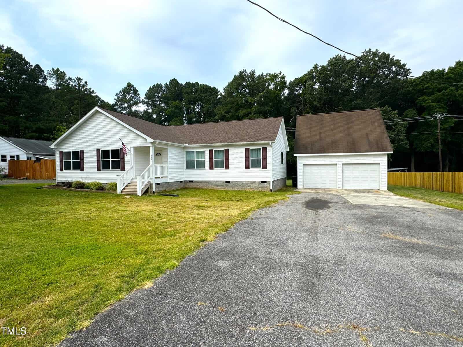 A one-story white house with a brown roof stands proudly at 603 12th Street in Butner. It features a front porch, a double garage, and a driveway leading to the street. The grassy yard showcases a U.S. flag on the porch, with trees adding charm in the background.