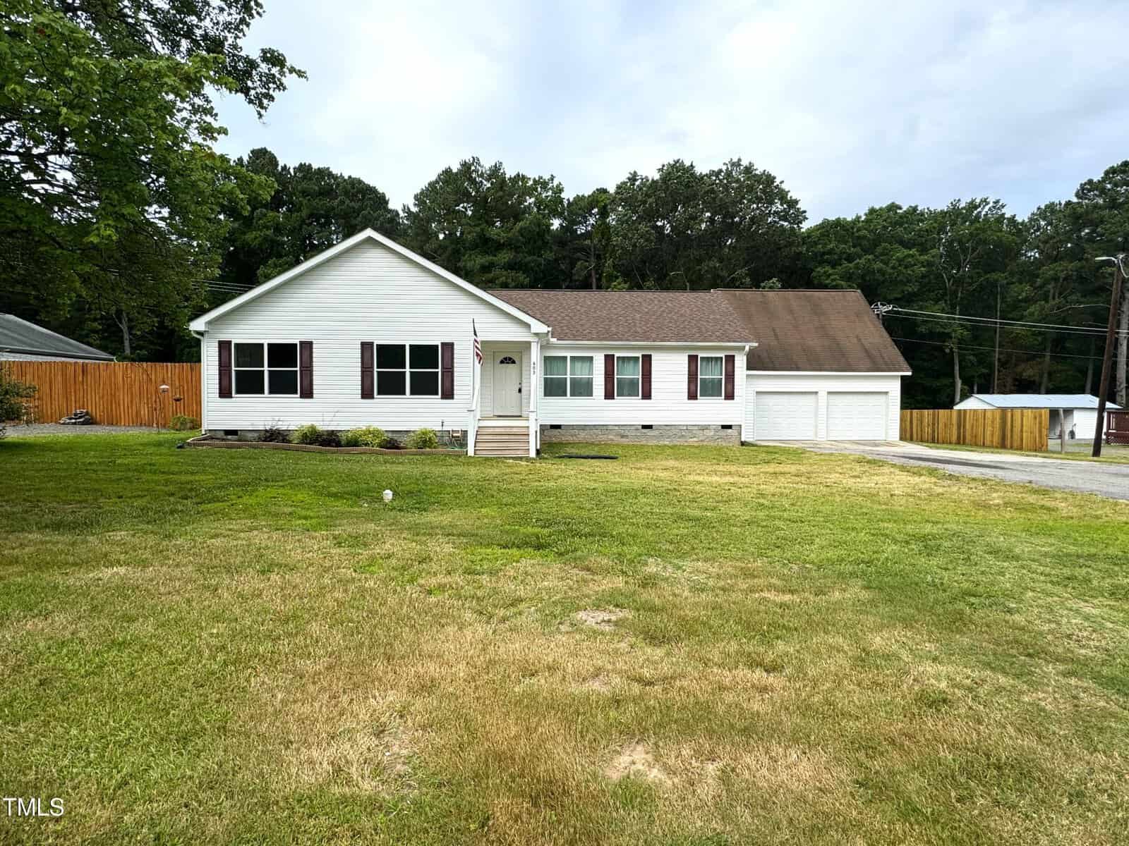 A white single-story house with a front porch and attached two-car garage. The yard is expansive and grassy, with a few trees in the background. A wooden fence lines part of the property.