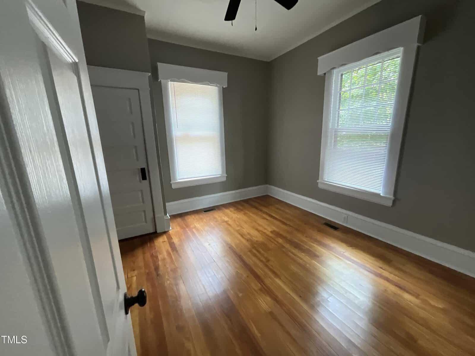 A room with hardwood floors, gray walls, and white trim, featuring two windows with blinds. A white door with black hardware is partially open, revealing another room. A ceiling fan is visible on the ceiling.