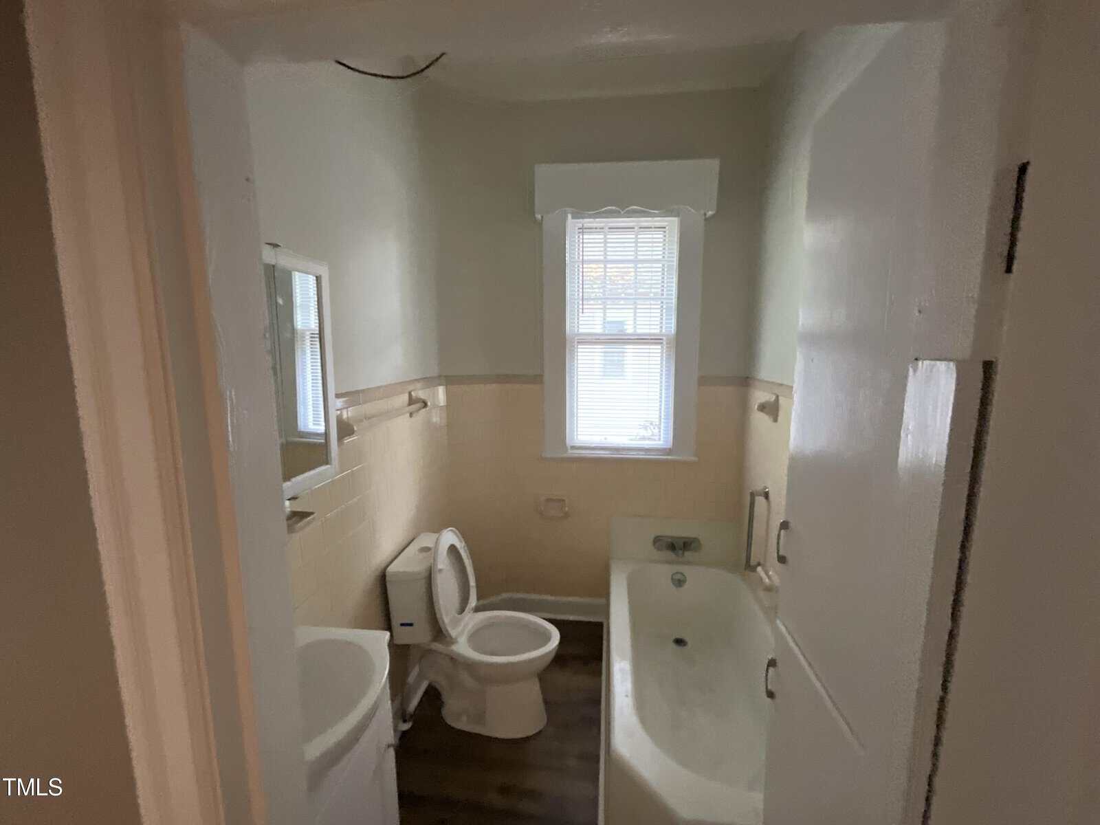 A small bathroom with beige tiled walls, a white toilet, sink, and bathtub. There is a window above the bathtub, allowing natural light to enter. The floor has dark wood-like vinyl flooring. A mirror is above the sink, and a towel bar is beside it.