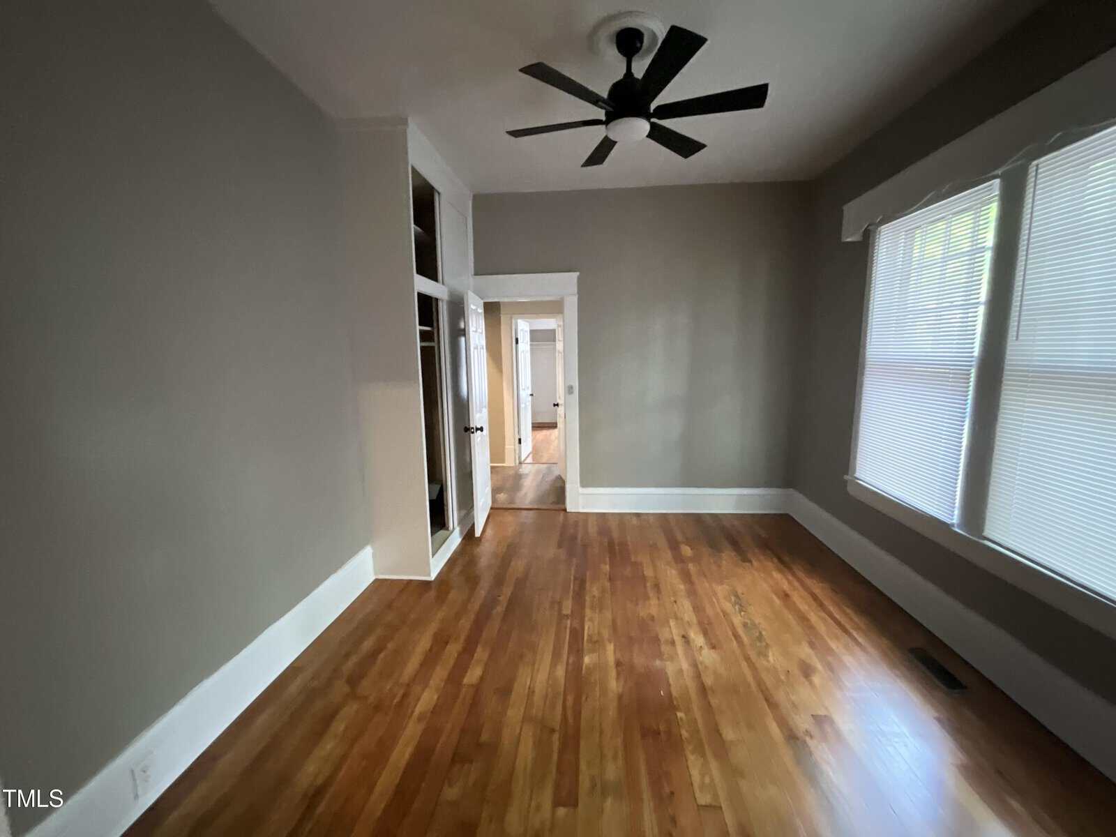 A room with wooden floors, gray walls, and white trim. There is a ceiling fan with a light in the center of the ceiling and large windows with blinds on one wall. The room has an open doorway leading to a hallway and a built-in shelving unit to the left of the doorway.