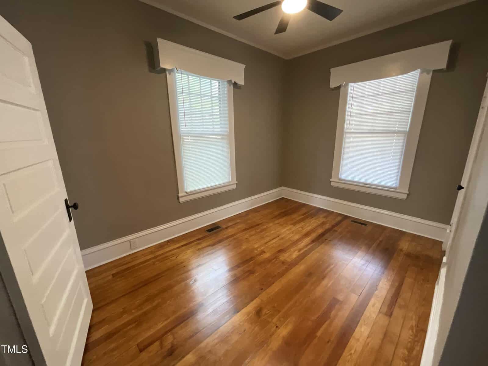 A small, empty room with hardwood floors, painted beige walls, two windows with blinds, a ceiling fan, and a white door on the left wall. The room is well-lit from natural light coming through the windows.