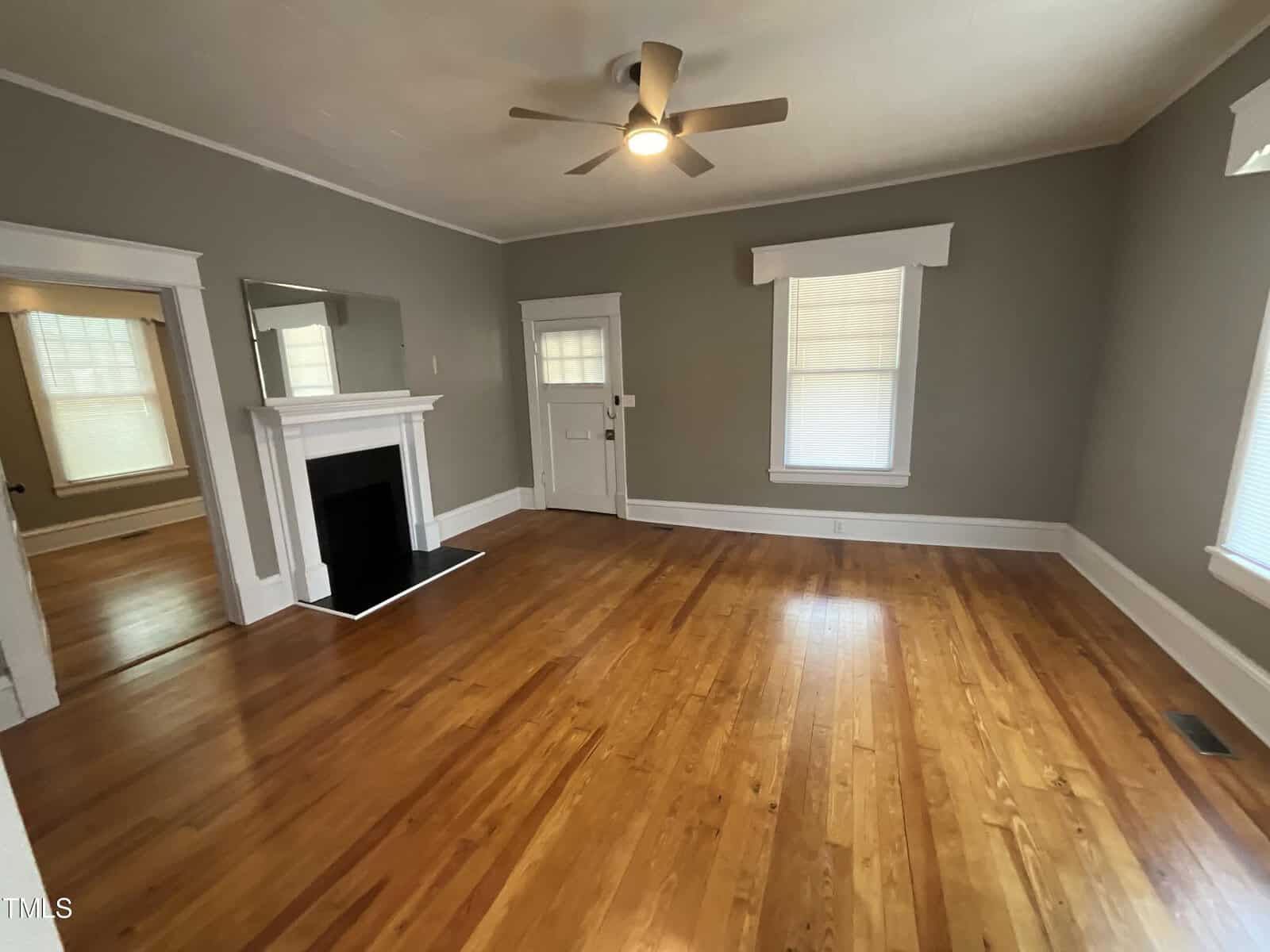 An empty living room with polished hardwood floors, gray walls, and white trim. The room has a ceiling fan, a fireplace with a mantle, and several windows with blinds. A door is located near the fireplace, and an open doorway leads to another room.