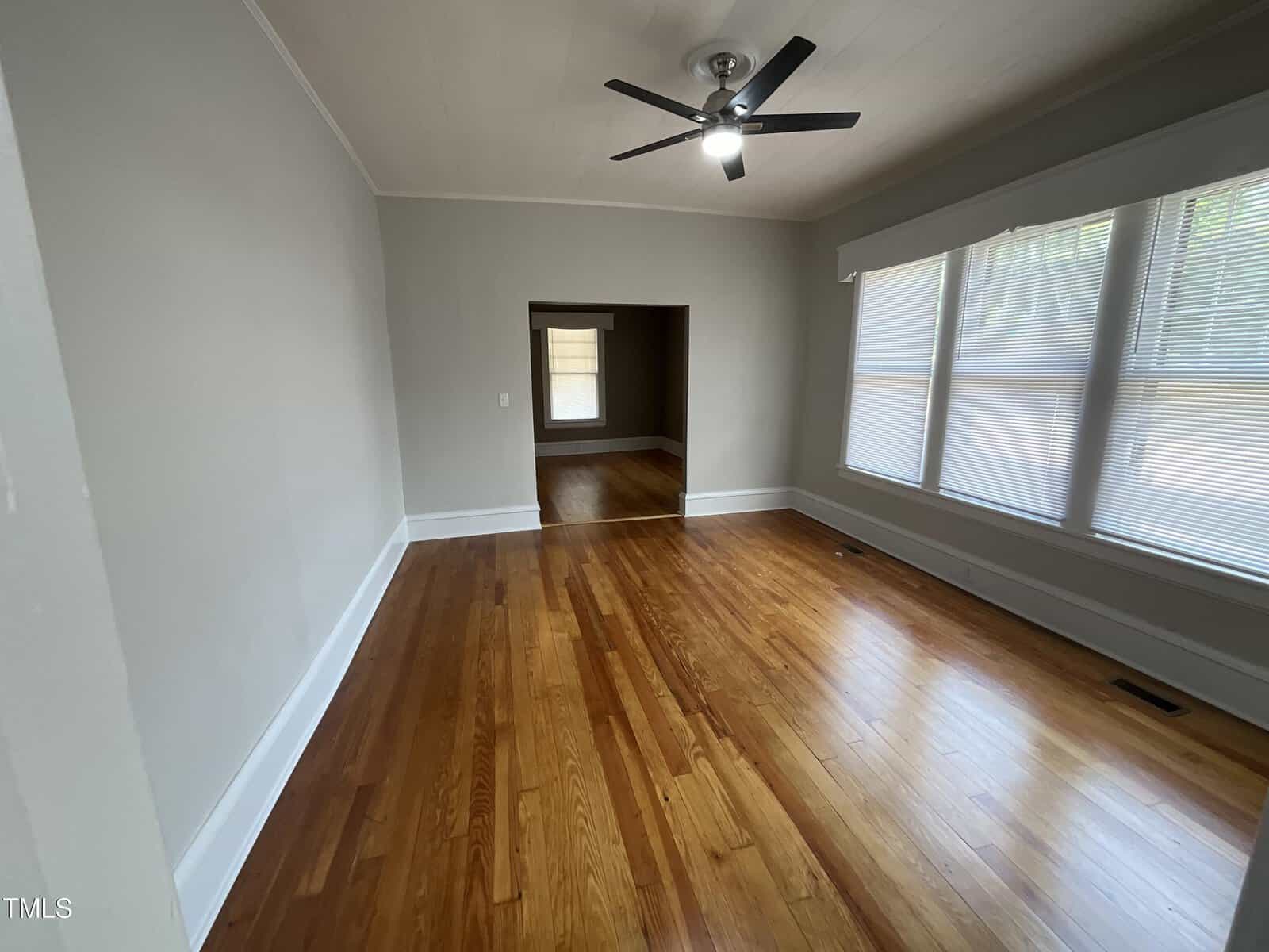 A spacious, empty room with polished hardwood floors and light gray walls. A ceiling fan with a light is centered on the ceiling. Large windows with blinds are on the right wall, and an open doorway leads to another room in the background.