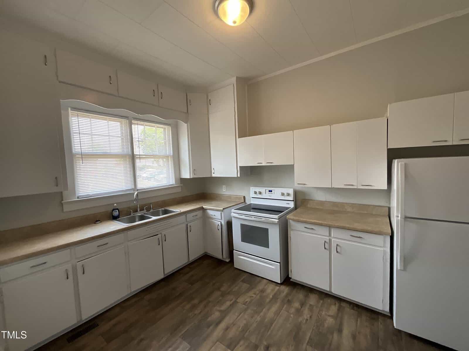 A kitchen with white cabinets, a beige countertop, a sink under a double window, and a white stove next to a refrigerator. The floor is dark wood, and the walls are painted light gray. The ceiling light fixture provides illumination.