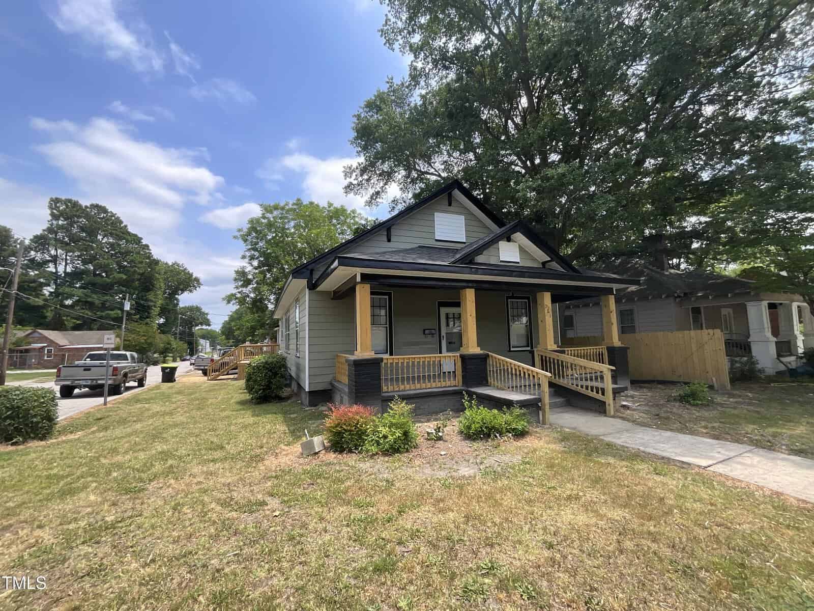 A single-story gray house with a dark gray roof sits on a corner lot at 701 S Williams Street, Henderson NC. It has a covered front porch with wooden pillars and a ramp leading up to the front door. The yard is mostly grass with a few small bushes, and large trees provide shade in the background.