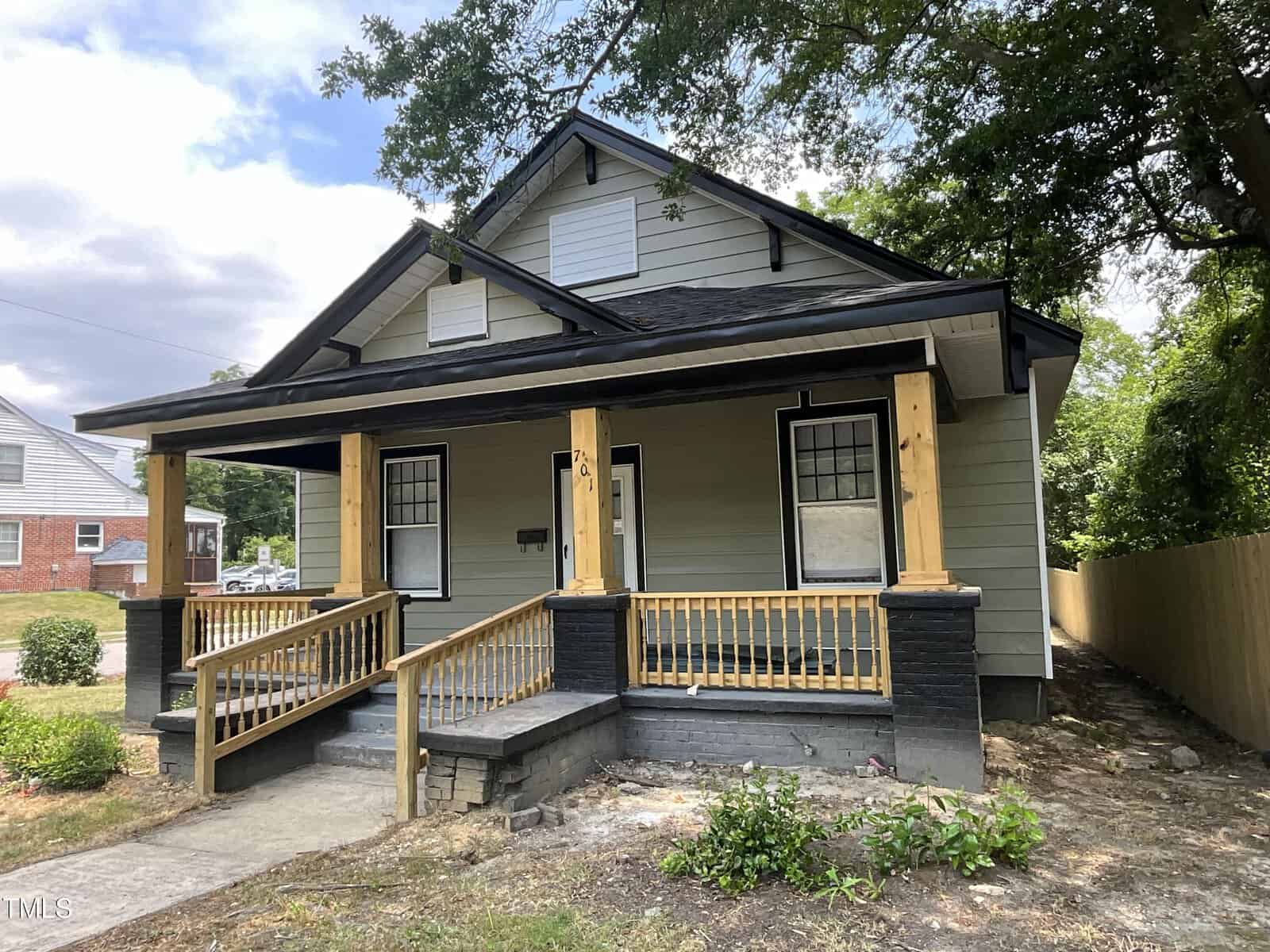 A small, single-story house with a covered front porch, light gray exterior, black trim, and a wooden railing. The front lawn has patches of grass and a concrete pathway leading to the porch. Trees and neighboring houses are visible in the background.
