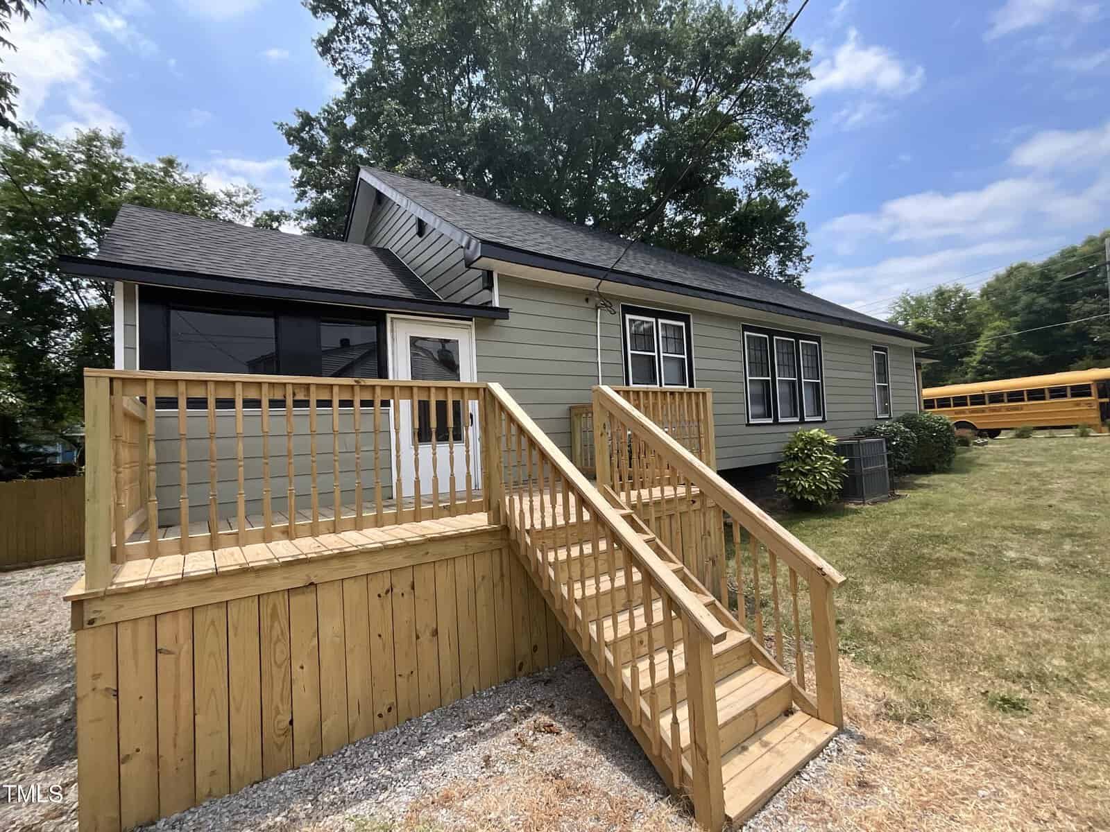 A single-story house with gray siding and white trim features a wooden deck with steps leading to the entrance. The house has several windows, a large tree in the background, and a yellow school bus partially visible in the driveway. The sky is partly cloudy.