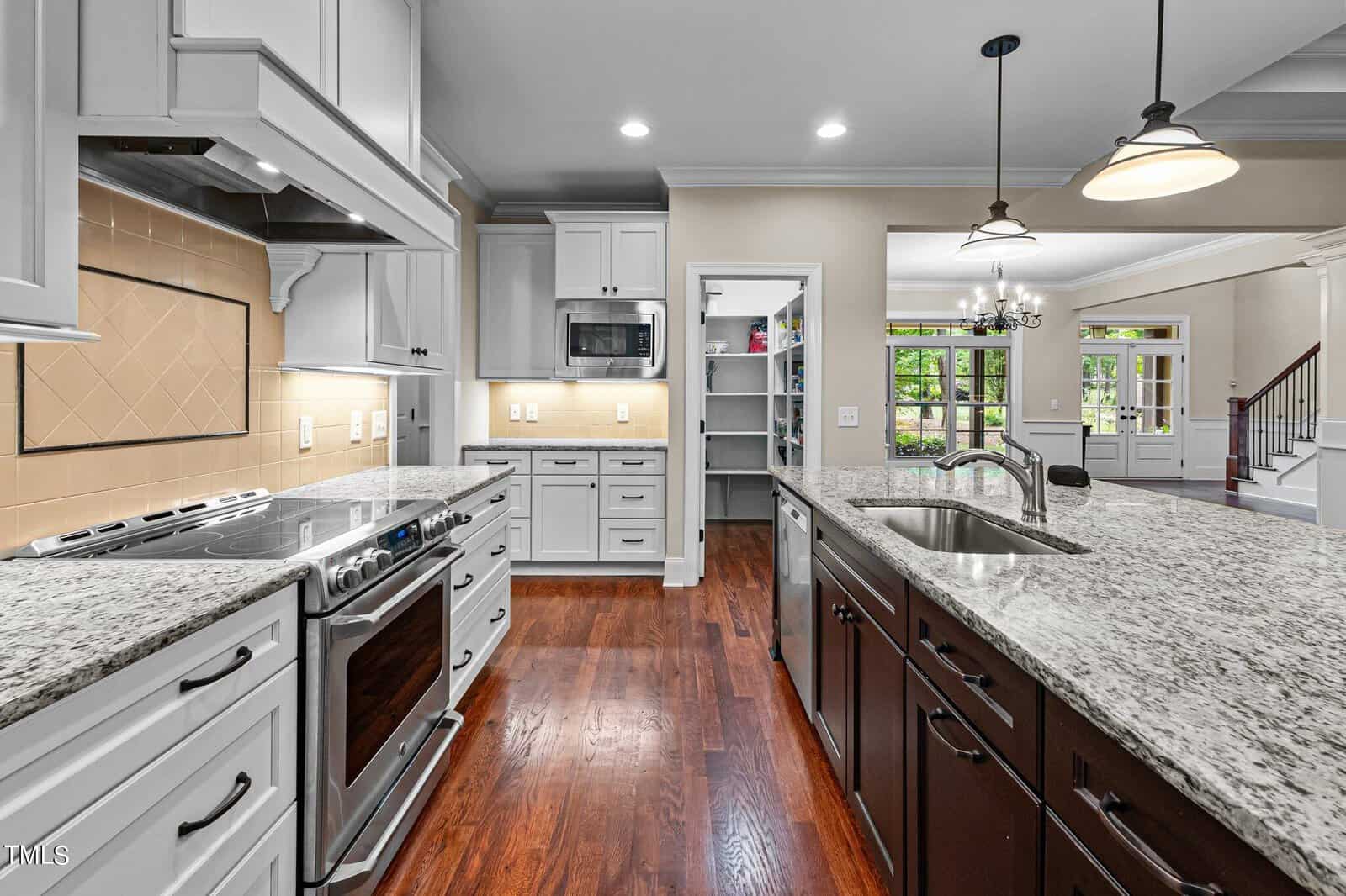 A modern kitchen with granite countertops, an island with a sink, white cabinetry, stainless steel appliances, and hardwood floors. A pantry with open door is visible. A dining area with a chandelier is seen in the background through an open doorway.