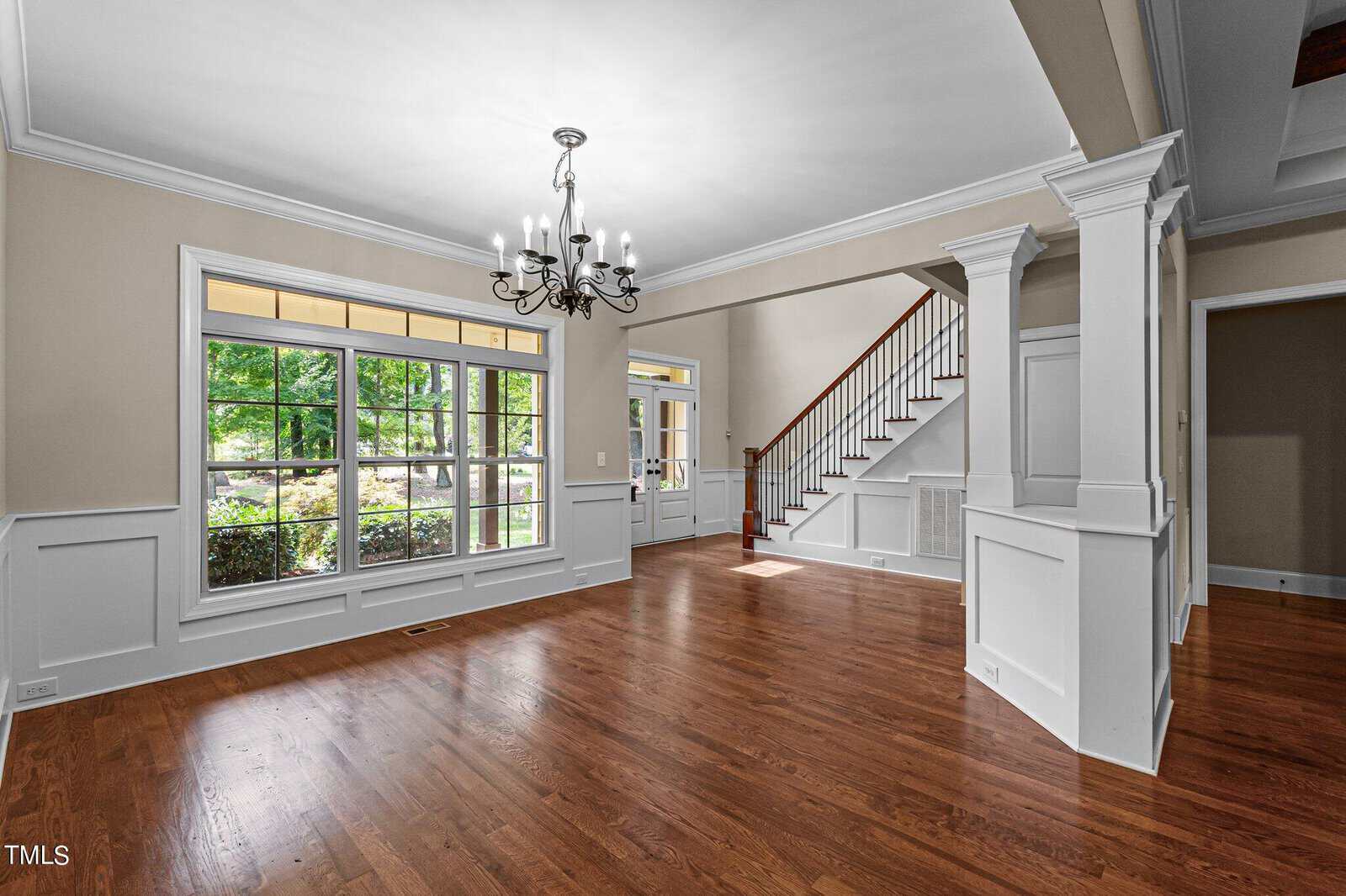 A spacious, empty room with hardwood floors and white wainscoting features a large window with a view of greenery. A grand staircase with wooden railings leads to the upper floor. A classic chandelier hangs from the ceiling, illuminating the room.