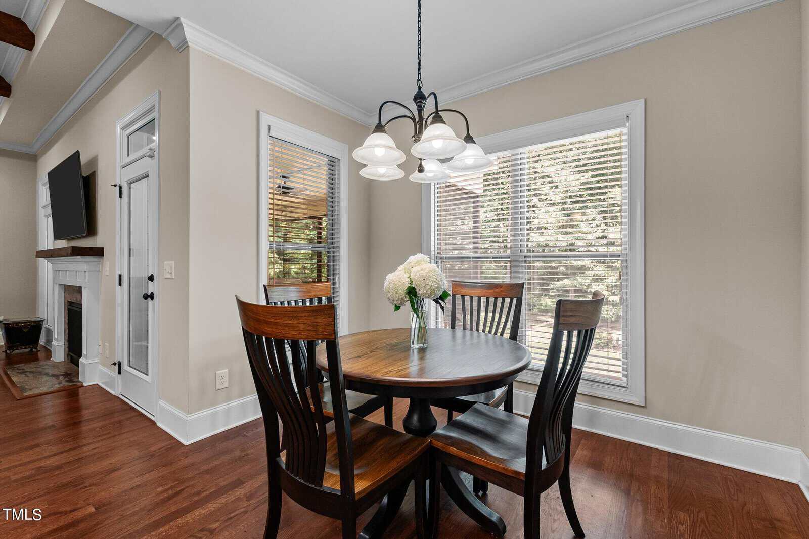 A cozy dining area with a round wooden table and four chairs, centered by a vase of white flowers. The room features hardwood floors, large windows with blinds, and a modern chandelier overhead. A TV and fireplace are visible in the adjacent room.