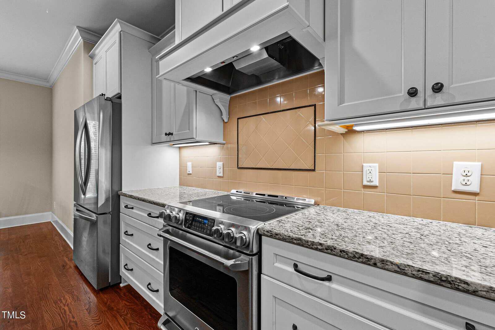 Modern kitchen with stainless steel appliances, including a refrigerator and oven. Granite countertops and beige tiled backsplash complement the white cabinetry. Warm wooden flooring adds contrast.