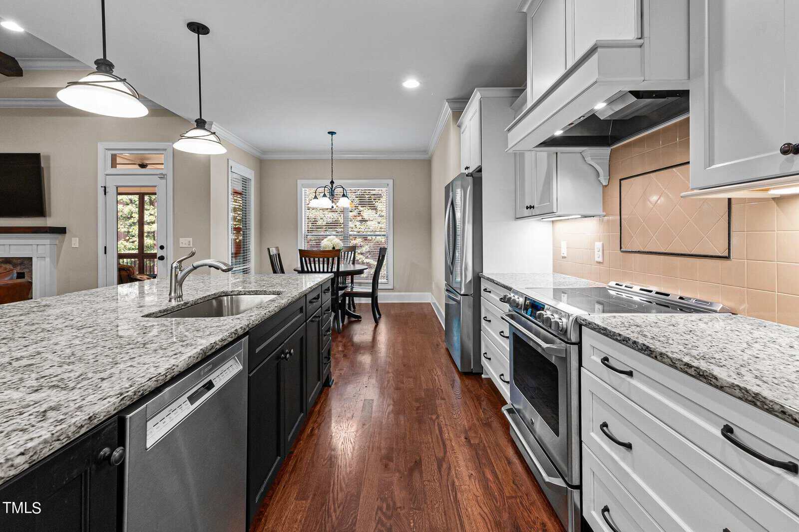 A modern kitchen features a long island with granite countertops and a built-in dishwasher. Pendant lights hang above. There's a stainless steel stove and refrigerator. The dining area in the background has a table, chairs, and a large window.