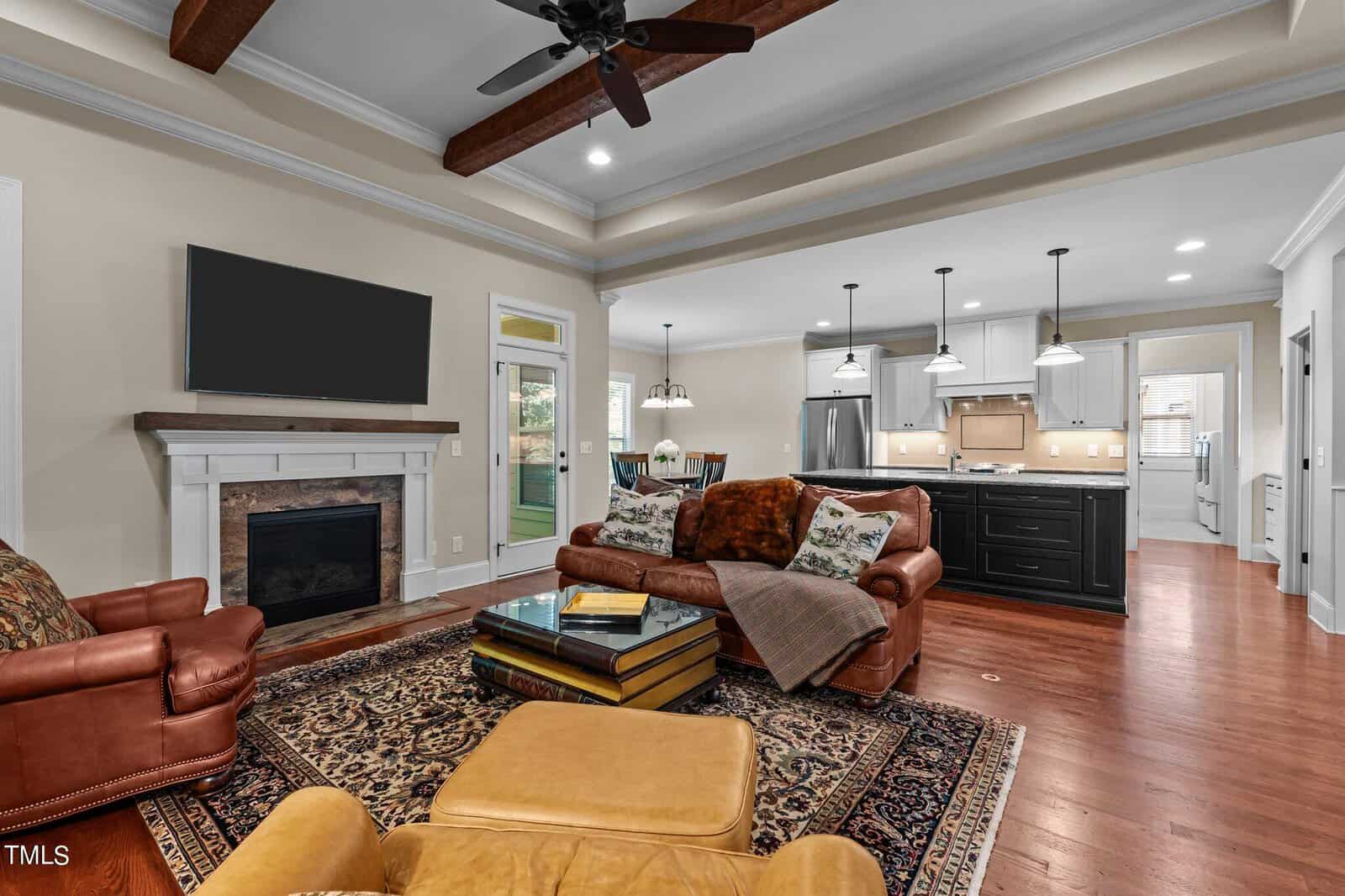 Spacious living room with leather armchairs, a large ottoman, and a patterned rug. A TV is mounted above the fireplace. The open kitchen features pendant lights, bar stools, and modern appliances. Wooden ceiling beams add a rustic touch.