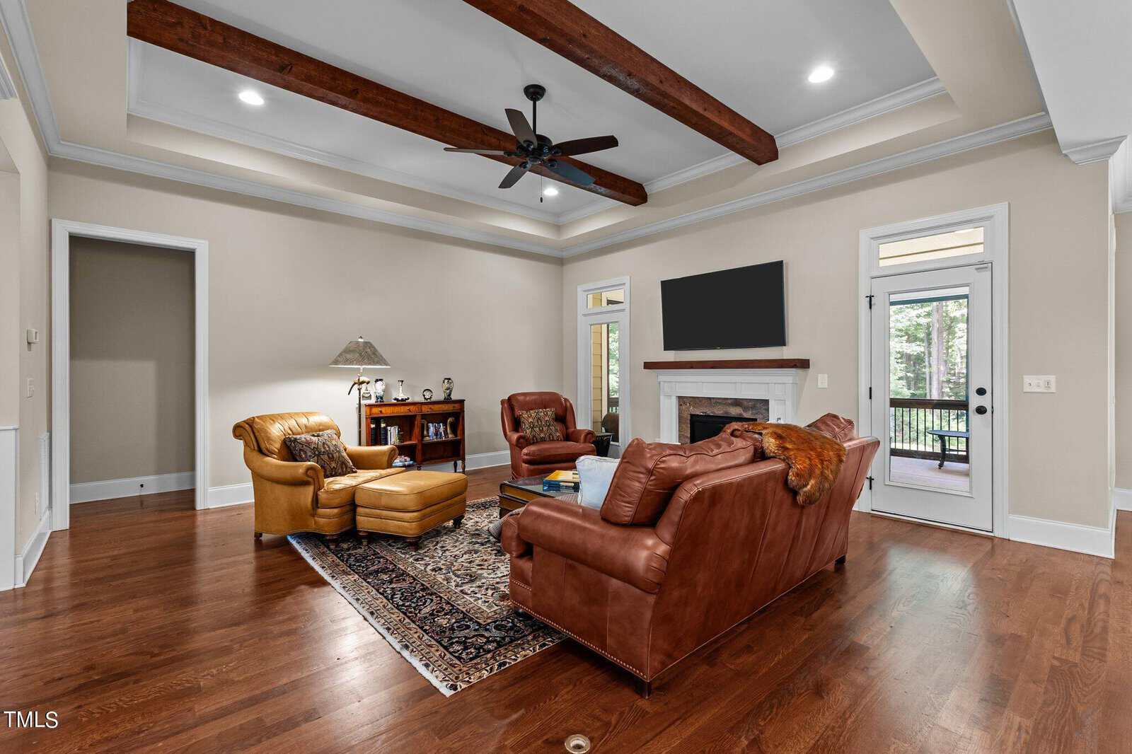 A cozy living room with a warm color scheme features a leather armchair and matching ottoman, a sofa, a wooden coffee table, and an area rug. The room has exposed wooden beams, a ceiling fan, and a large TV mounted above a fireplace. A door opens to a patio.