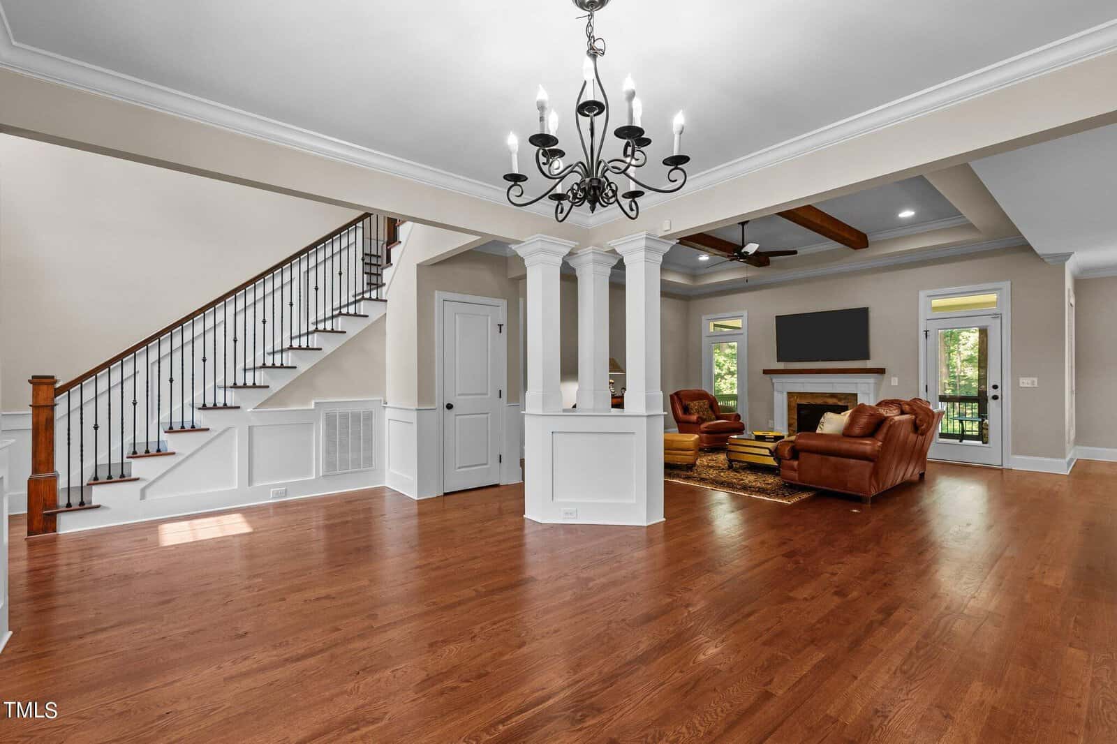 A spacious living room with hardwood floors, a chandelier, and a fireplace. Leather sofas are arranged around the fireplace, and tall windows provide natural light. A staircase with wooden railing is on the left, and the room features elegant columns.