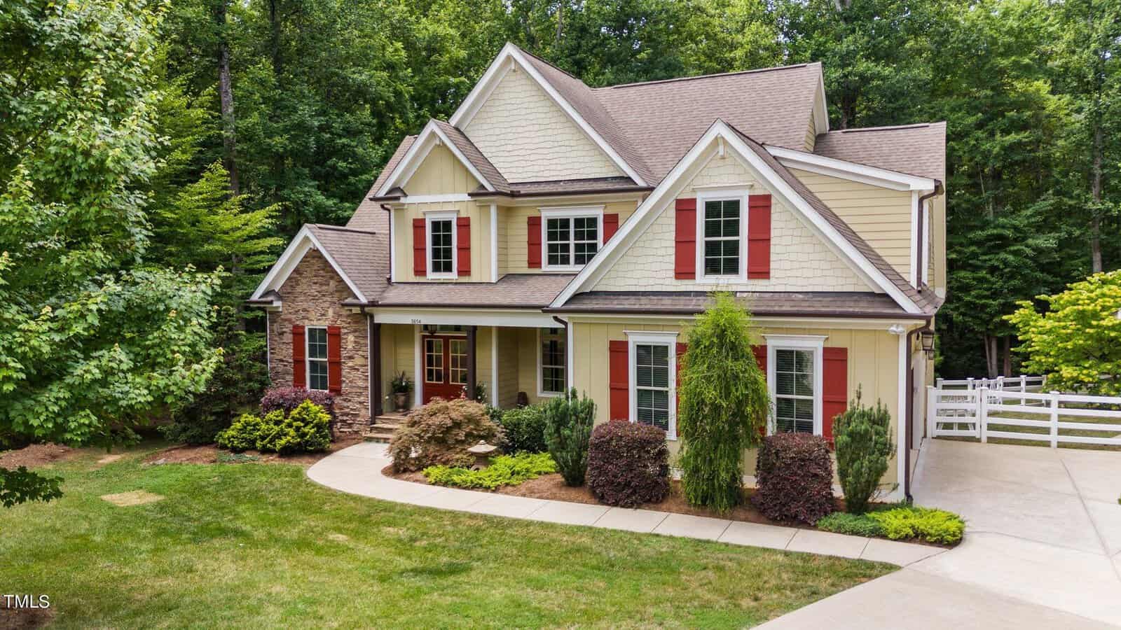 A two-story yellow house with red shutters and a stone accent section. The house is surrounded by green trees and bushes, with a neatly manicured lawn and a white driveway. There is a small garden visible at the front right corner.