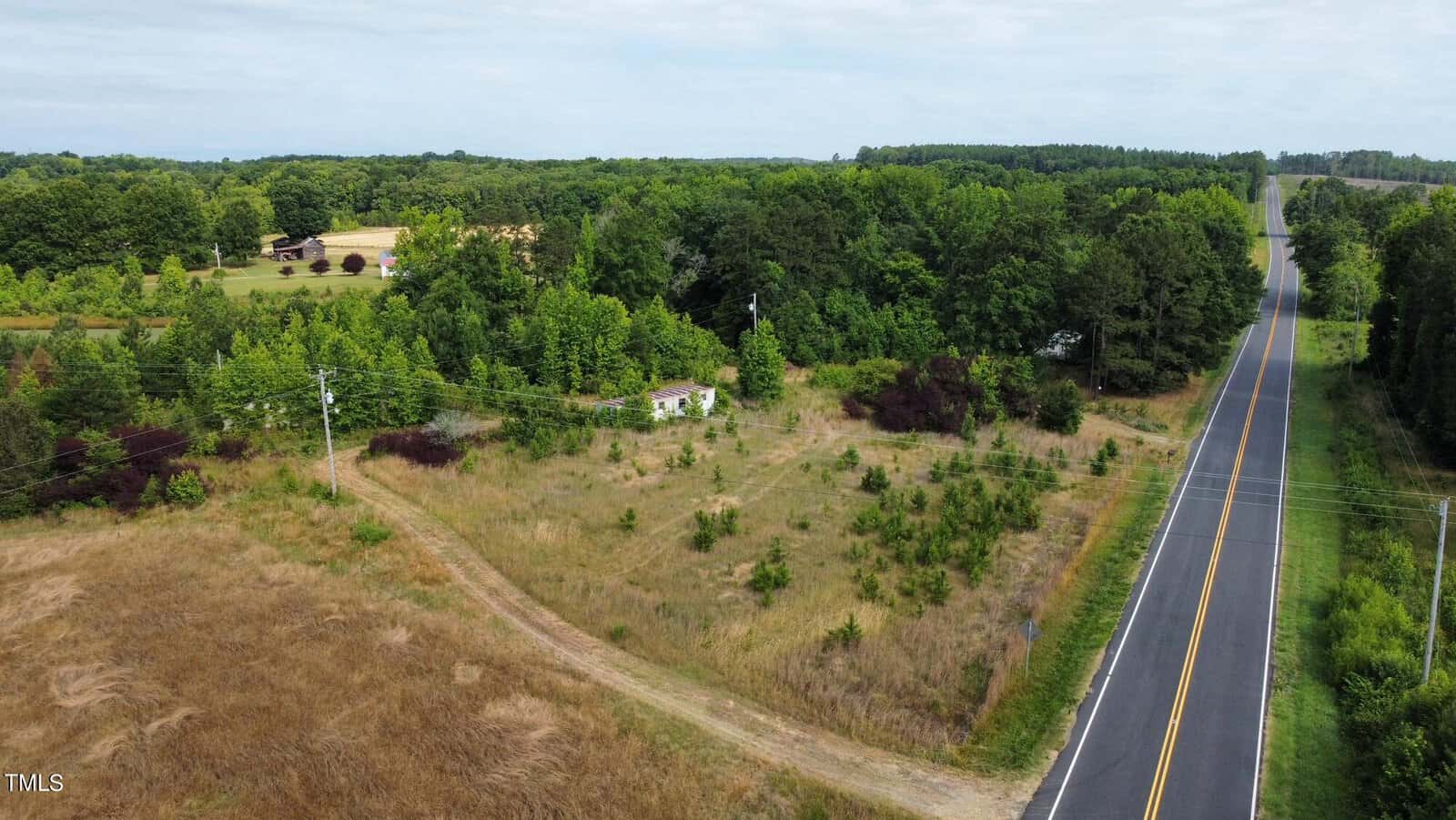 Aerial view of a rural landscape with a straight road running alongside a small, grassy field. A few scattered trees and shrubs are visible near the field, with a dense forest in the background and a house in the distance.