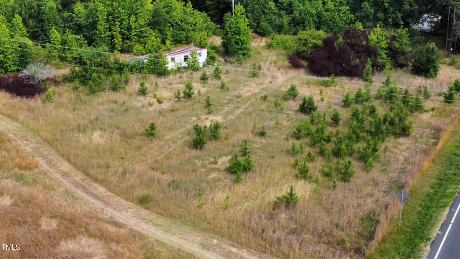 Aerial view of a grassy field with scattered small trees. A small, abandoned white building with a partially collapsed roof is situated near the center. Power lines cross above, and dense green trees border the area.