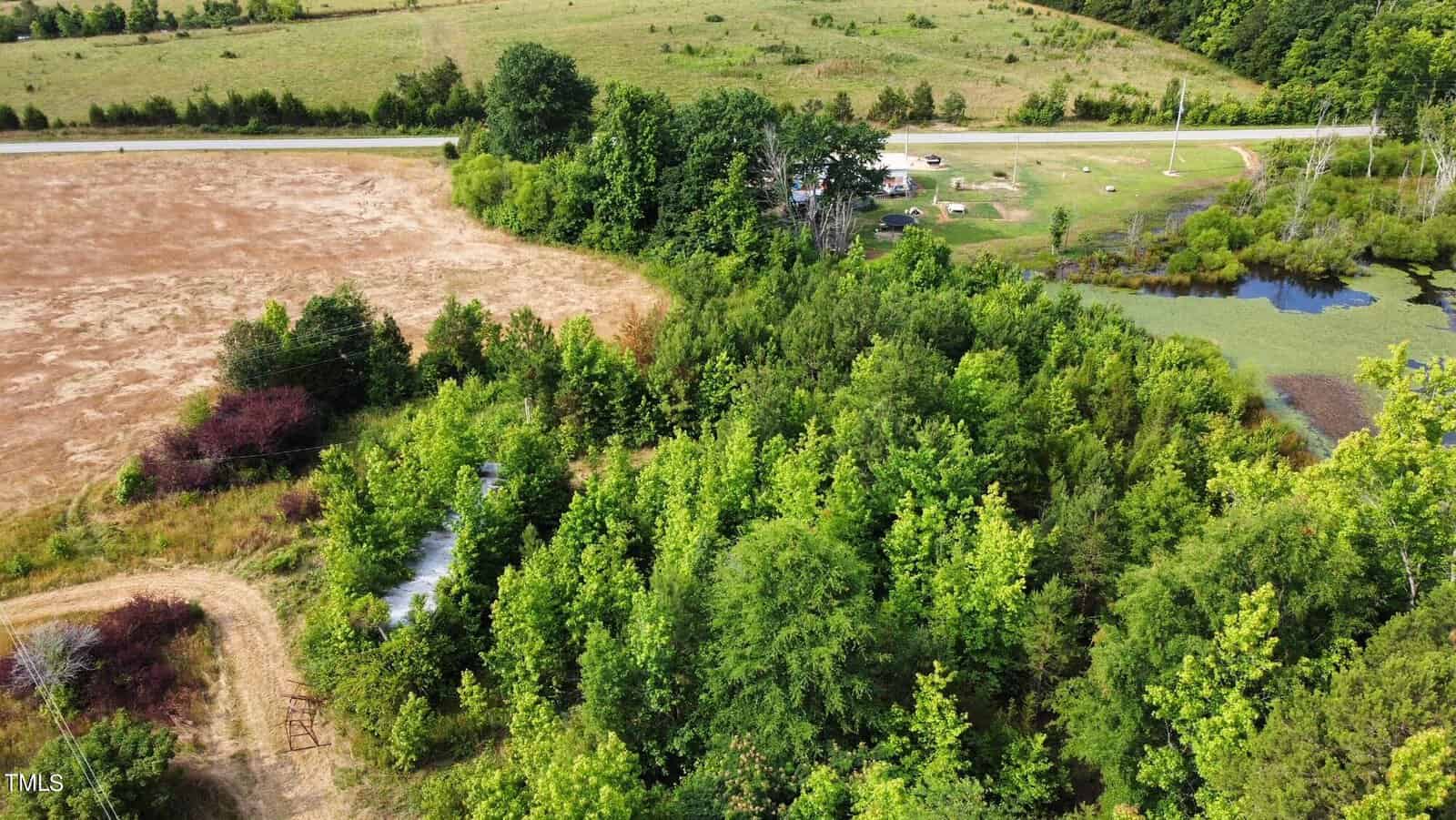 Aerial view of a rural landscape with a mix of lush green trees and dry fields. A road runs through the background, with a few buildings and a pond to the right. The area is surrounded by dense vegetation and open farmland.