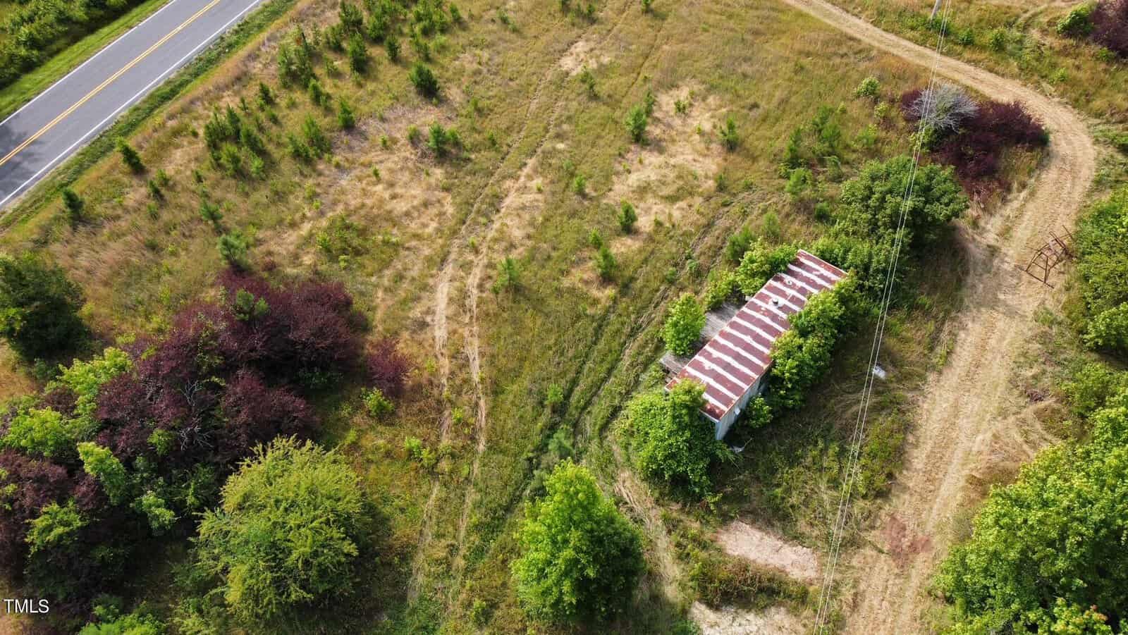 Aerial view of an overgrown rural landscape with a small building featuring a rusted metal roof surrounded by trees. A narrow dirt path leads to the structure, and a paved road is visible on the left side of the image.
