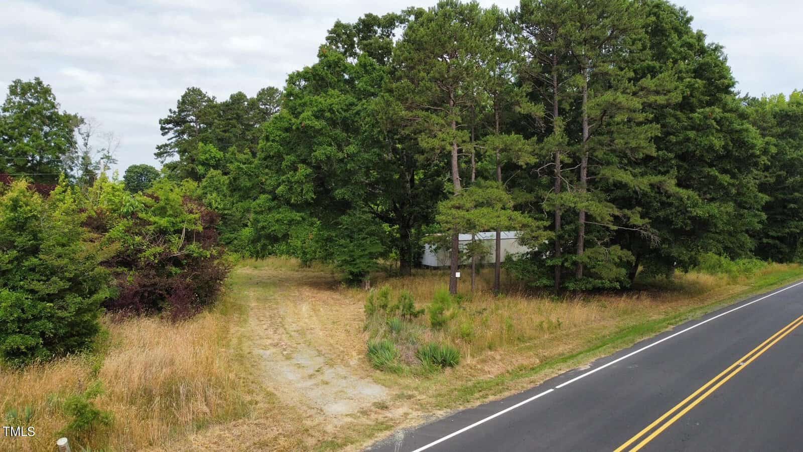 A rural scene featuring a small white building partially obscured by trees, next to a paved road. The landscape includes lush green foliage and patches of dry grass. A narrow dirt path leads from the road into the wooded area.
