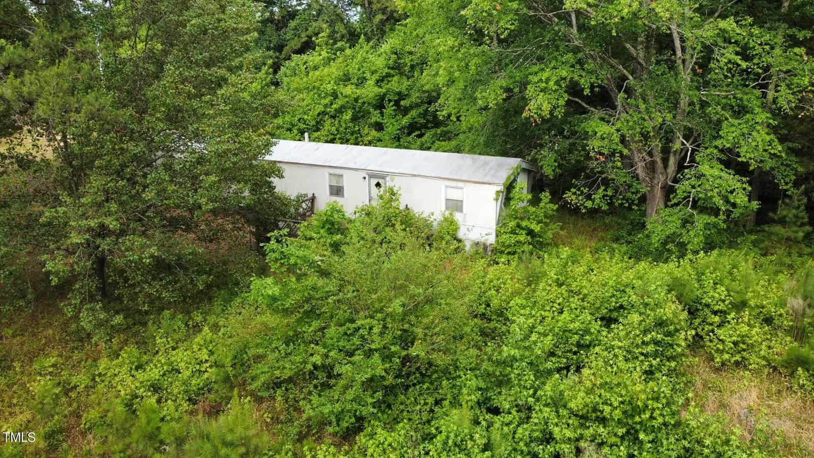 A white mobile home nestled amidst dense green foliage and trees. The setting appears to be a secluded, forested area, with lush vegetation surrounding the structure.