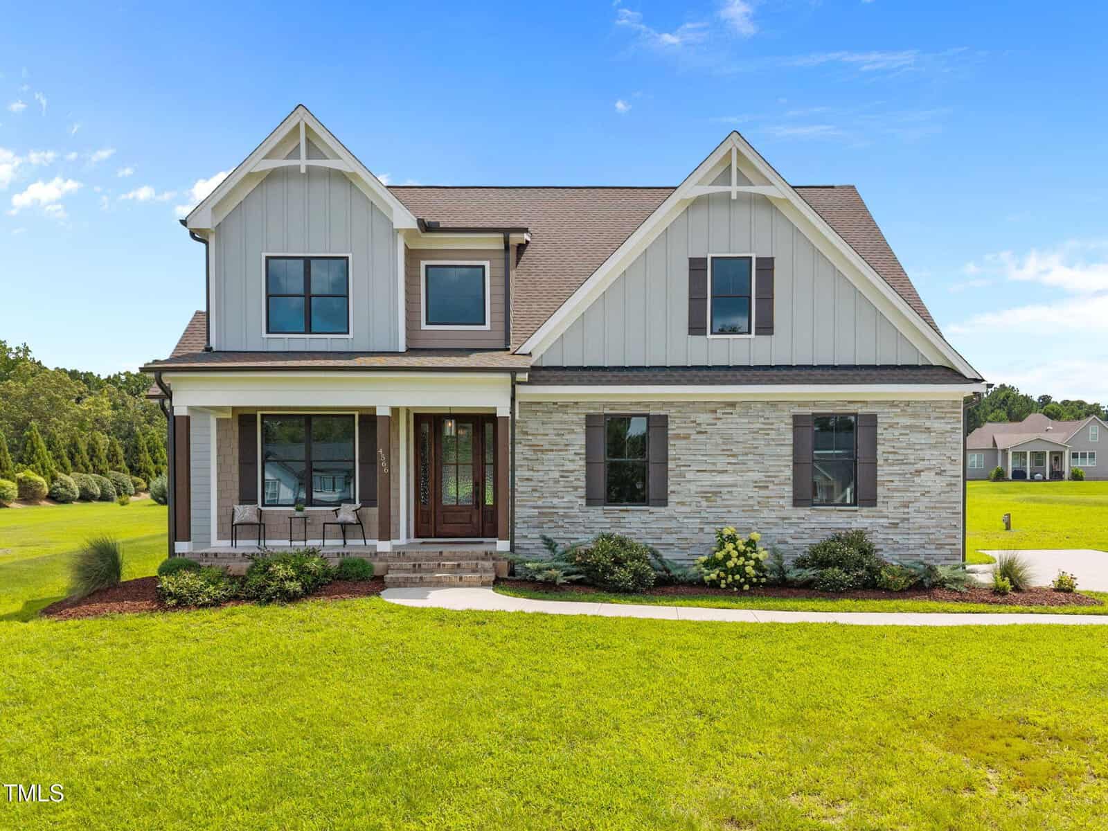 A modern two-story house with a gabled roof, light gray siding, and stone accents. The front has a covered porch with chairs, landscaped shrubs, and a large green lawn under a blue sky.