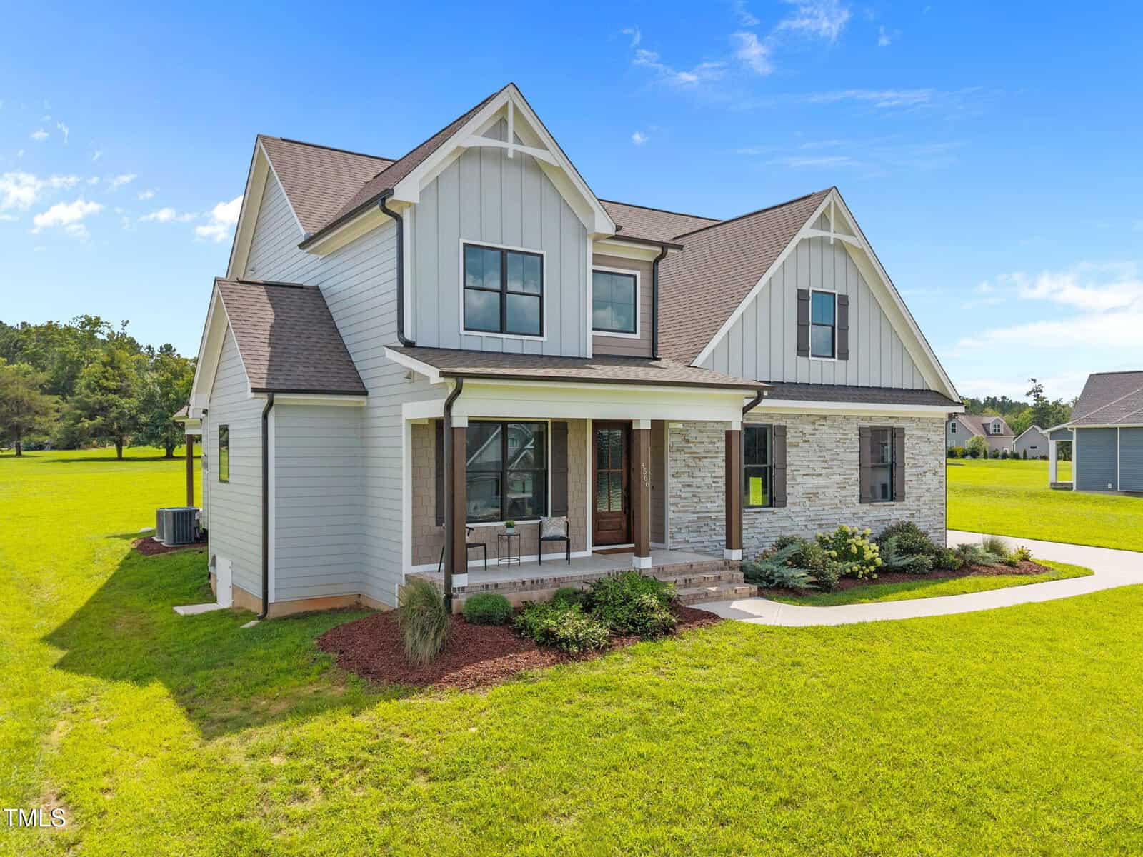 A two-story modern farmhouse with white siding, stone accents, and a covered front porch, situated on a green lawn under a blue sky with scattered clouds.