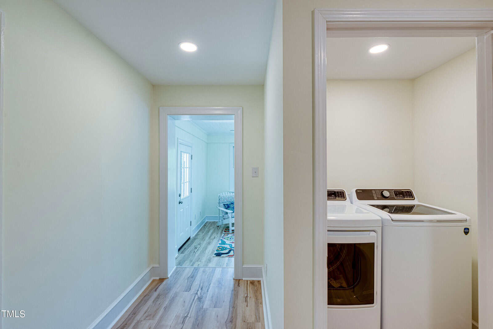 A hallway with light-colored walls and wood flooring leads to a laundry area with a washer and dryer on the right. Natural light enters from a door at the end of the hallway, and a small table is visible in the adjoining room.