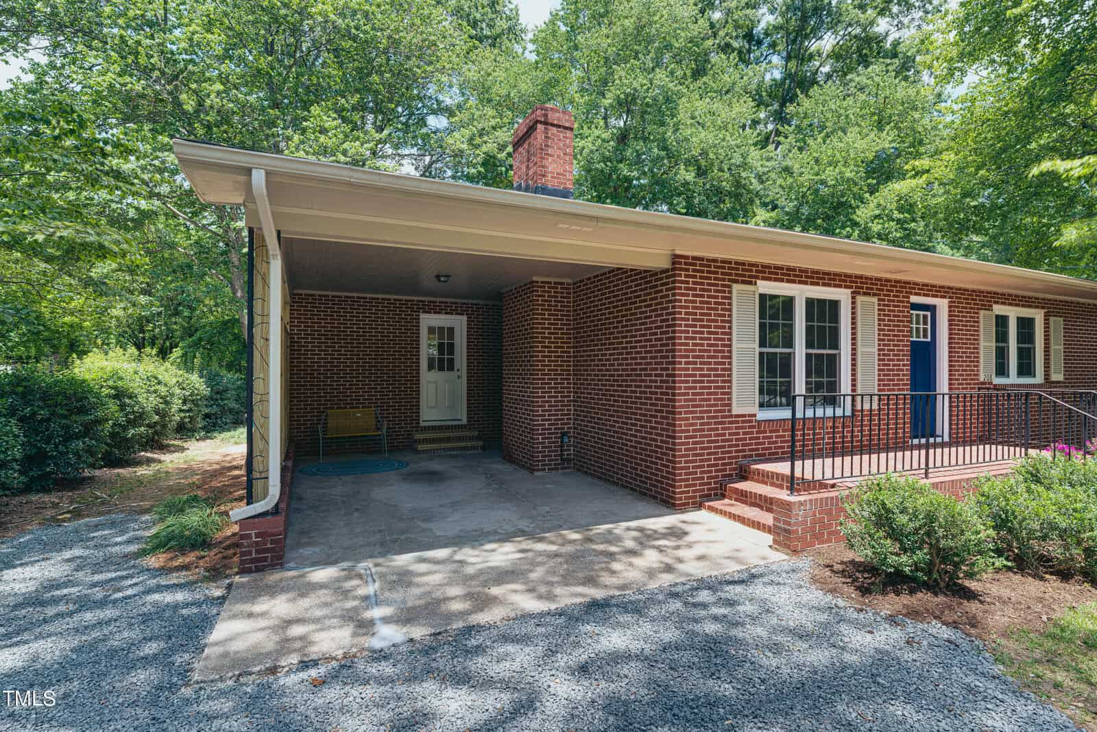 A brick house with a covered porch, featuring a white door and a blue door. It is surrounded by trees and shrubs, with a gravel driveway leading to the porch. A bright, sunny day highlights the greenery.