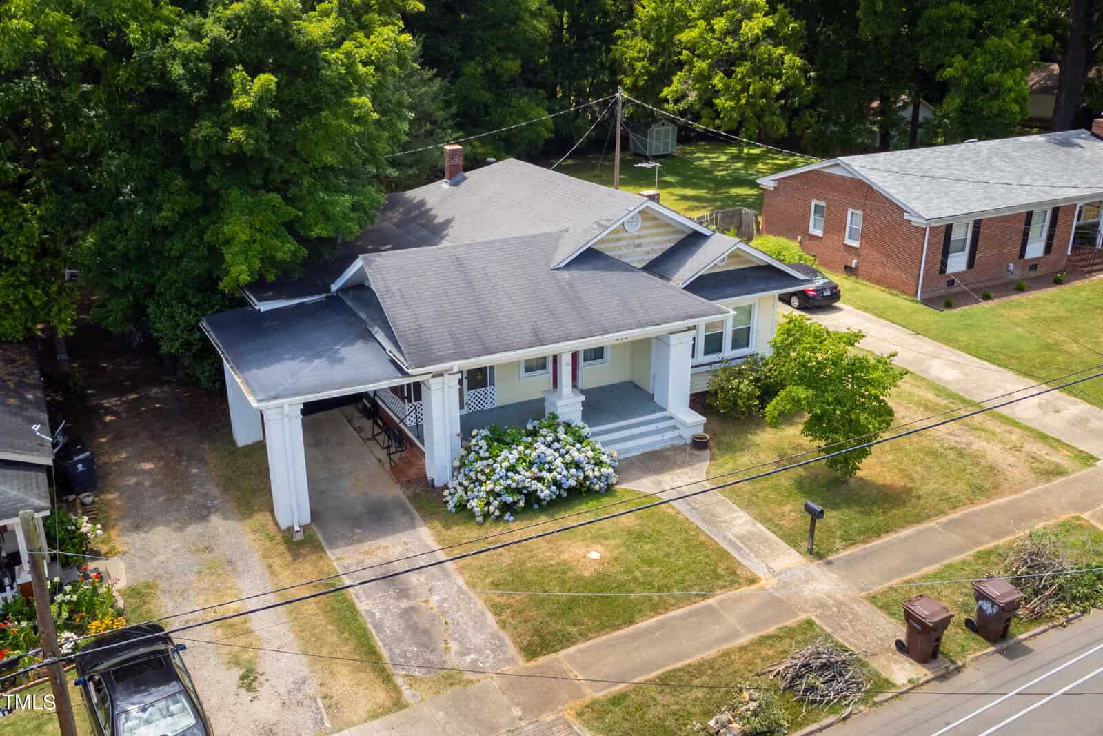 Aerial view of a single-story house with a gray roof and white exterior. The house has a covered porch, a driveway on the left, and blooming bushes in front. It's surrounded by green lawns and neighboring houses, with trees in the background.