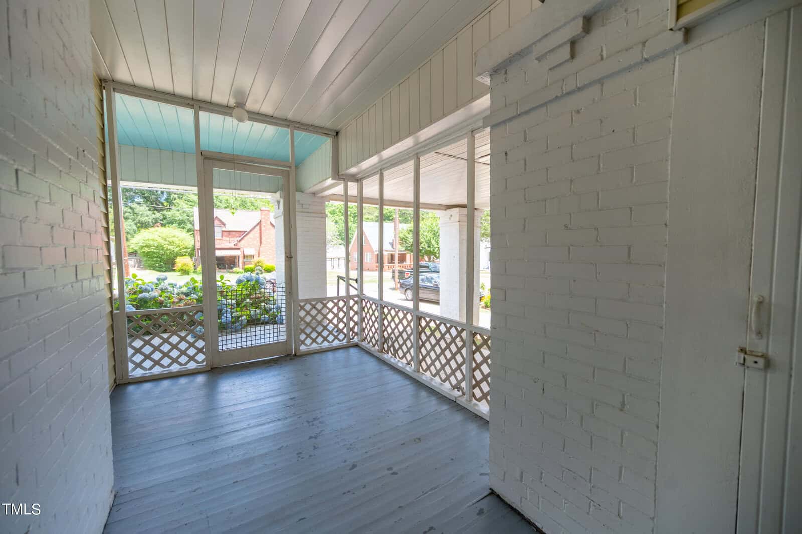 A bright, enclosed porch with white brick walls and a blue ceiling. The space features a wooden floor and lattice railing. Through the windows, a view of a garden with flowering bushes and a residential neighborhood can be seen.