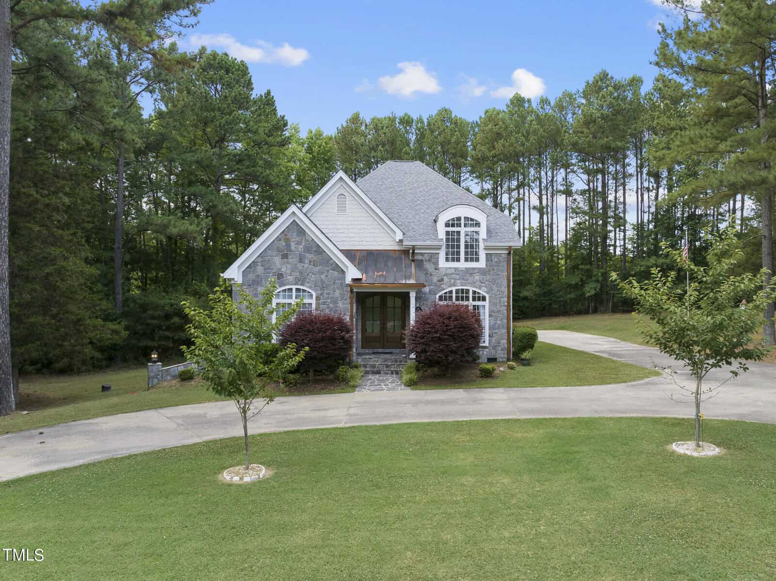 A two-story stone house at 55 Clare Drive, Henderson NC, with a light gray shingled roof is surrounded by tall trees. The house features large windows and a wooden balcony above the front porch. A circular driveway with small trees and neatly trimmed grass leads to the front entrance.