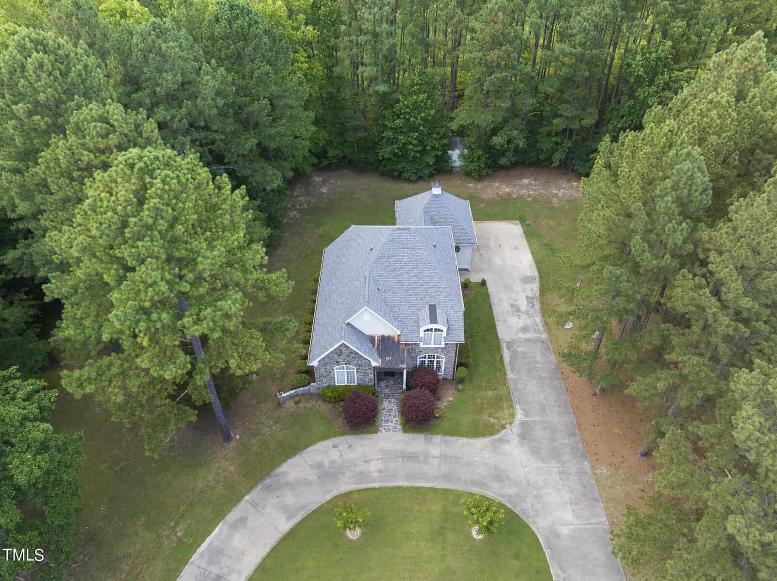 Aerial view of a single-story brick house surrounded by trees on all sides. The house is situated on a large, curved driveway with a well-maintained lawn. The driveway forms a loop in front of the house, providing ample space for parking.