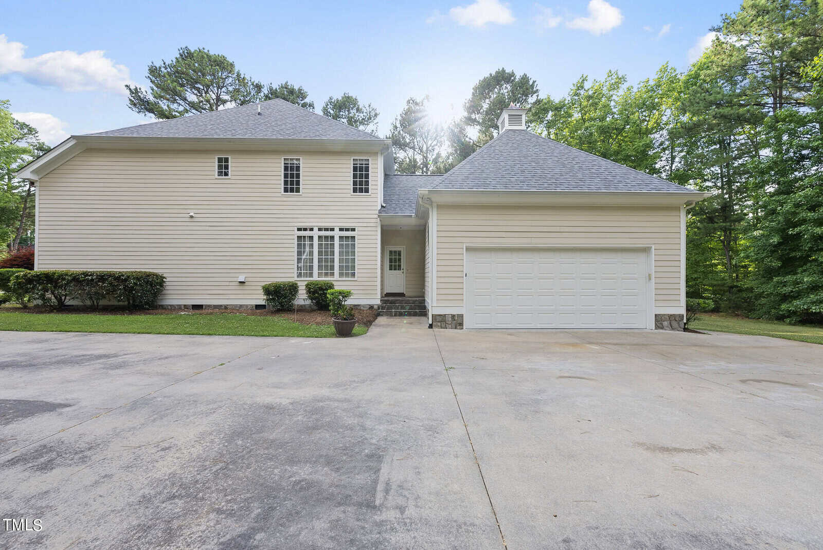 A two-story, light-colored house with a gray roof, attached two-car garage, and a spacious driveway. The house is surrounded by green trees and bushes. The sun is peeking through the trees, creating a serene and bright atmosphere.