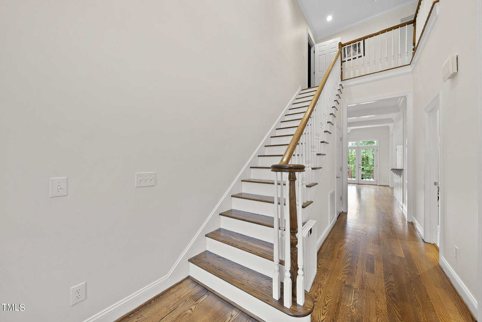 A bright, spacious entryway with wooden floors and a staircase with white risers on the left. The hallway extends towards the back with doorways to additional rooms. The walls are painted white, and there is a railing on the upper floor overlooking the entry.
