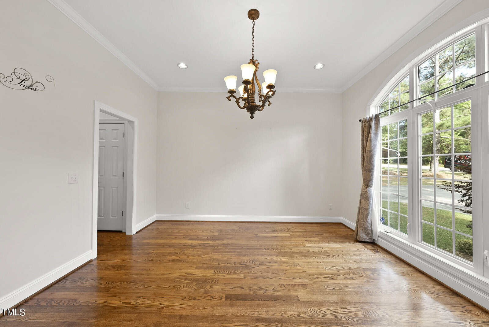 A bright, unfurnished room with polished hardwood floors, cream-colored walls, a large arched window with curtains, and a classic chandelier hanging from the ceiling. There is a door on the left side of the room. Natural light fills the space.