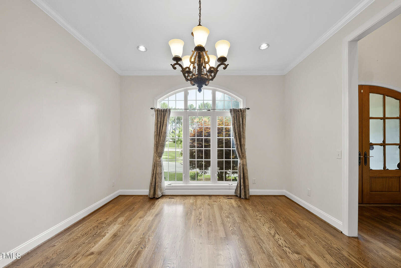 A bright, empty room with white walls and hardwood flooring. There's a large arched window with curtains and a view of a green, leafy backyard. An elegant chandelier hangs from the ceiling, illuminating the space. A wooden door is visible to the right.